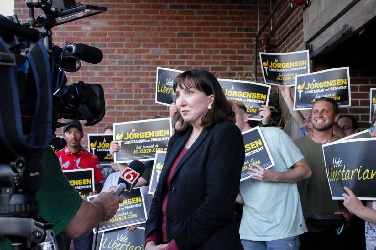 Jo Jorgensen being interviewed by TV crew with her supporters holding her poster behind her. None of the are wearing mask.